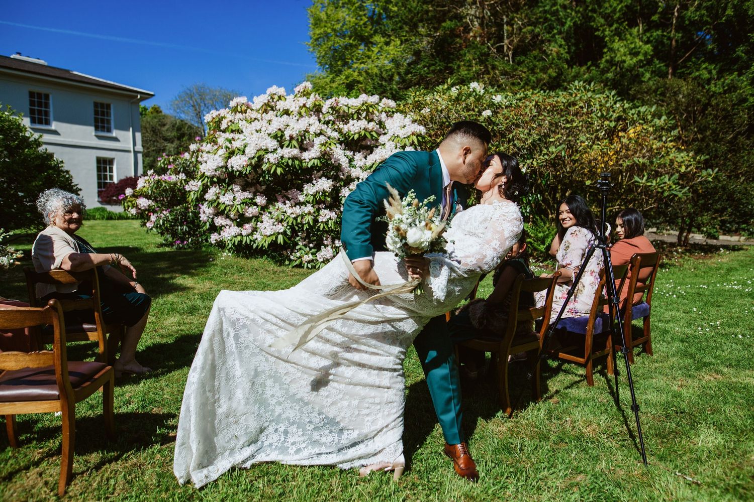 Five wedding reception chairs and decorative items are arranged outdoors on a lush green lawn with blooming flowers.