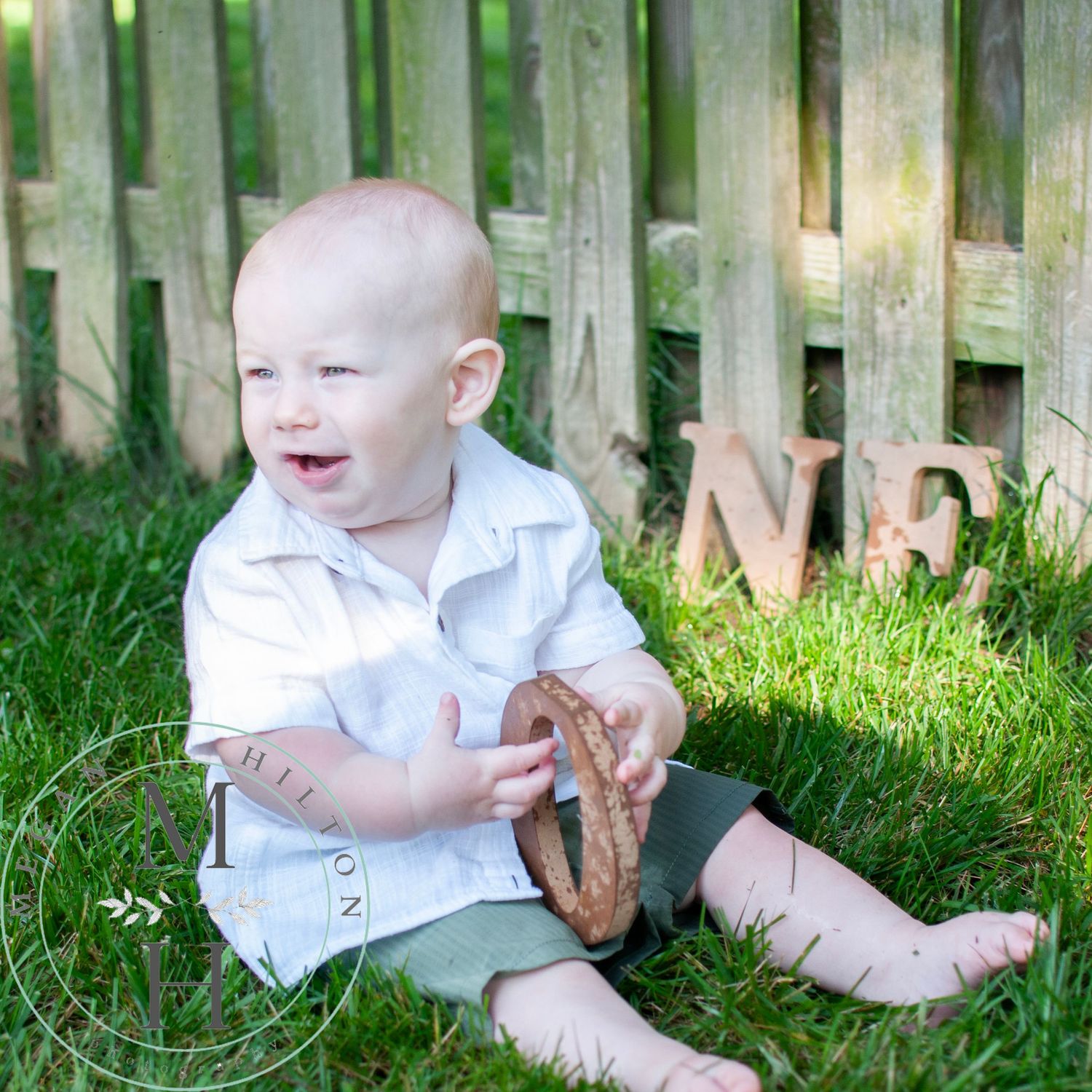 Small child in white shirt sitting on grass by wooden fence on sunny day laughs while holding a wooden O for ONE.