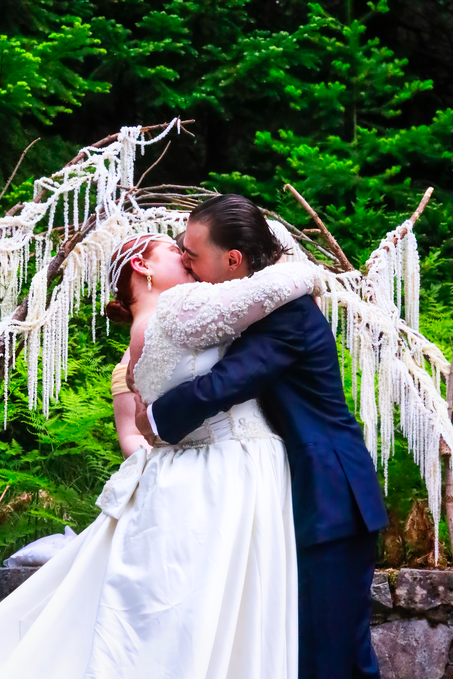 Two people share a romantic kiss under a decorative white arch with hanging crystals against a lush green forest backdrop.