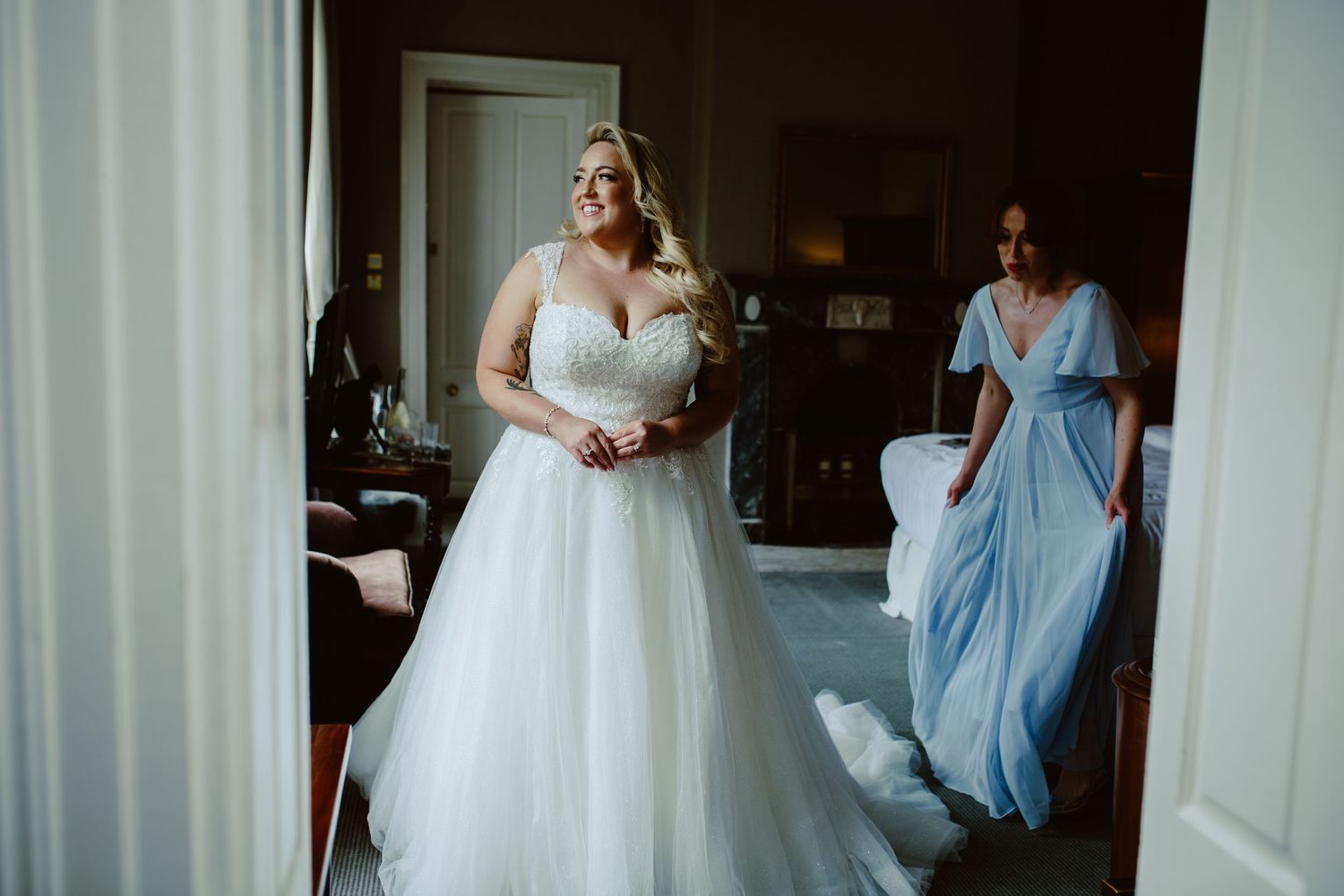 A bride in a traditional white ball gown prepares for her wedding day while an attendant in blue assists nearby.
