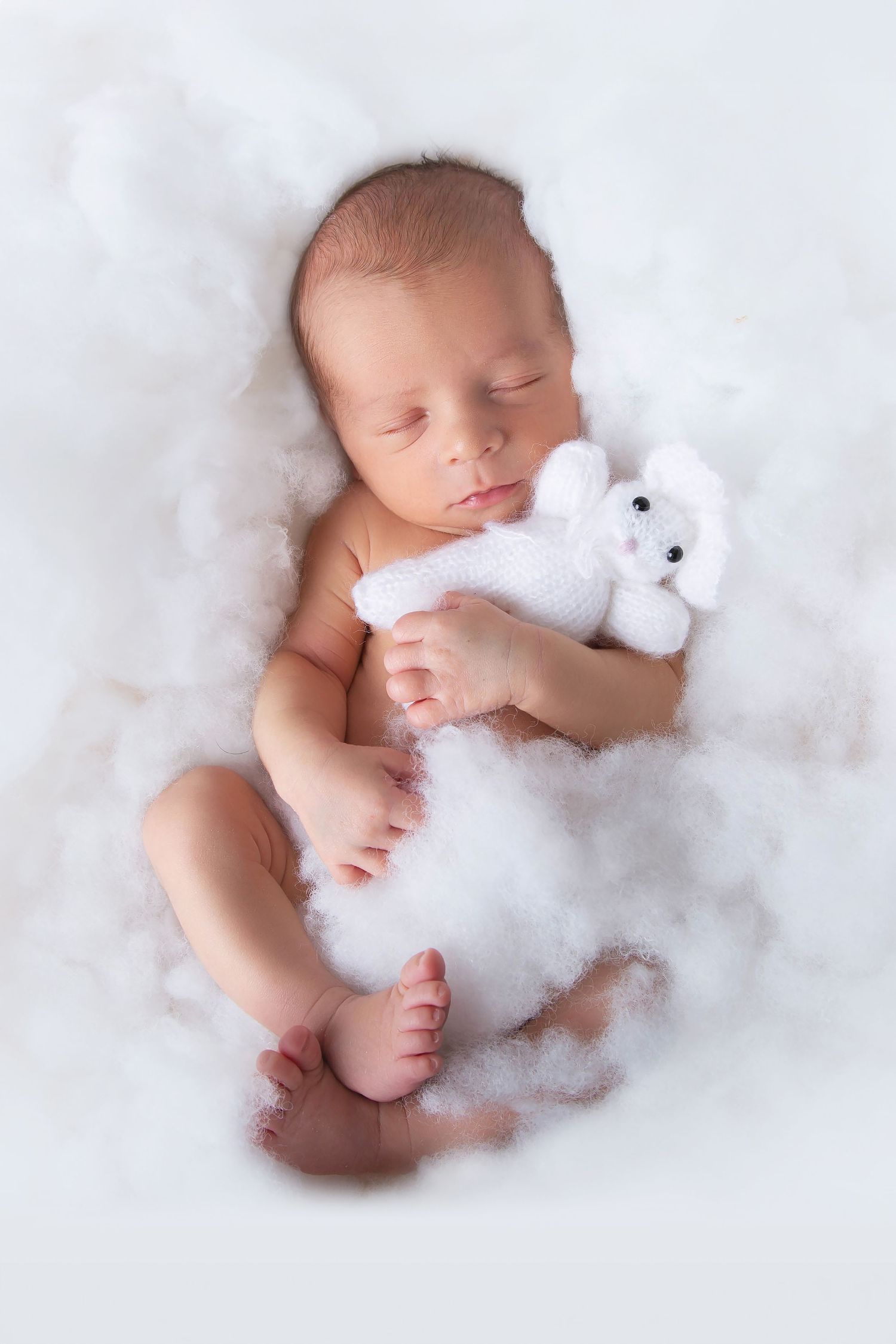 A peaceful newborn sleeping on fluffy white bedding while cuddling a small white teddy bear.