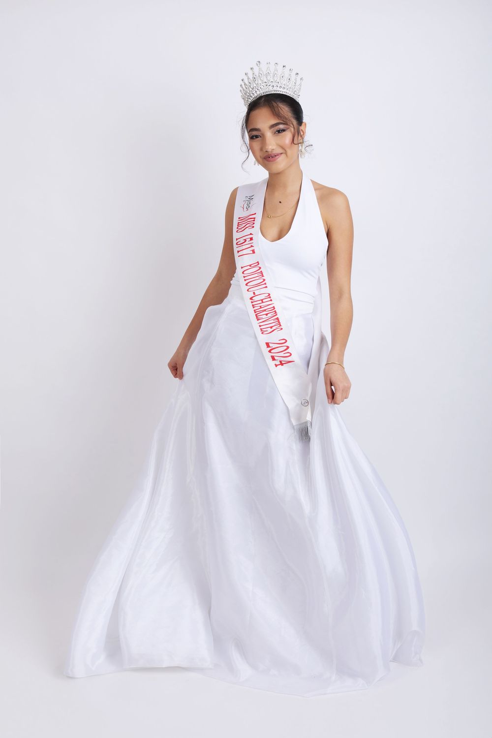 Person in flowing white ball gown wearing sash and crown poses elegantly against light background.