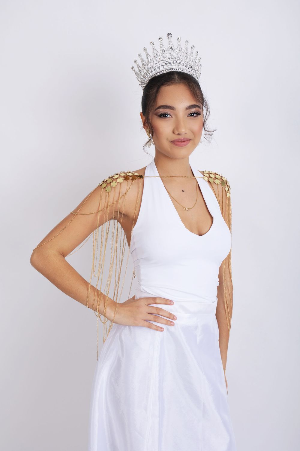 A pageant contestant in a white halter dress and crystal crown with gold shoulder tassels poses against a light background.