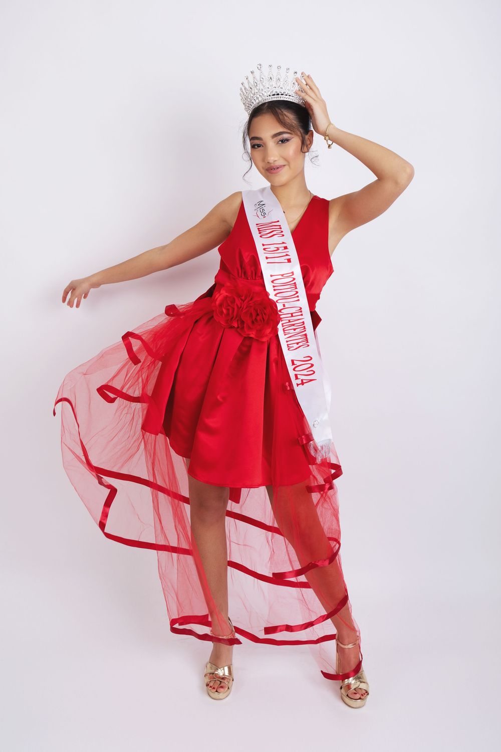 A pageant contestant in a flowing red gown and white sash poses gracefully against a light background.
