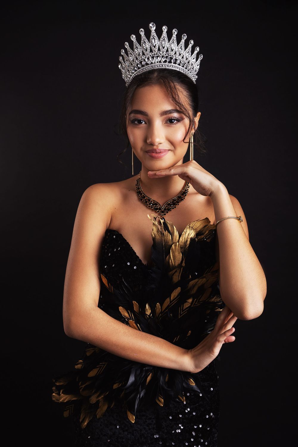A pageant contestant poses in a black strapless gown adorned with feathers and a sparkling crown against dark backdrop.