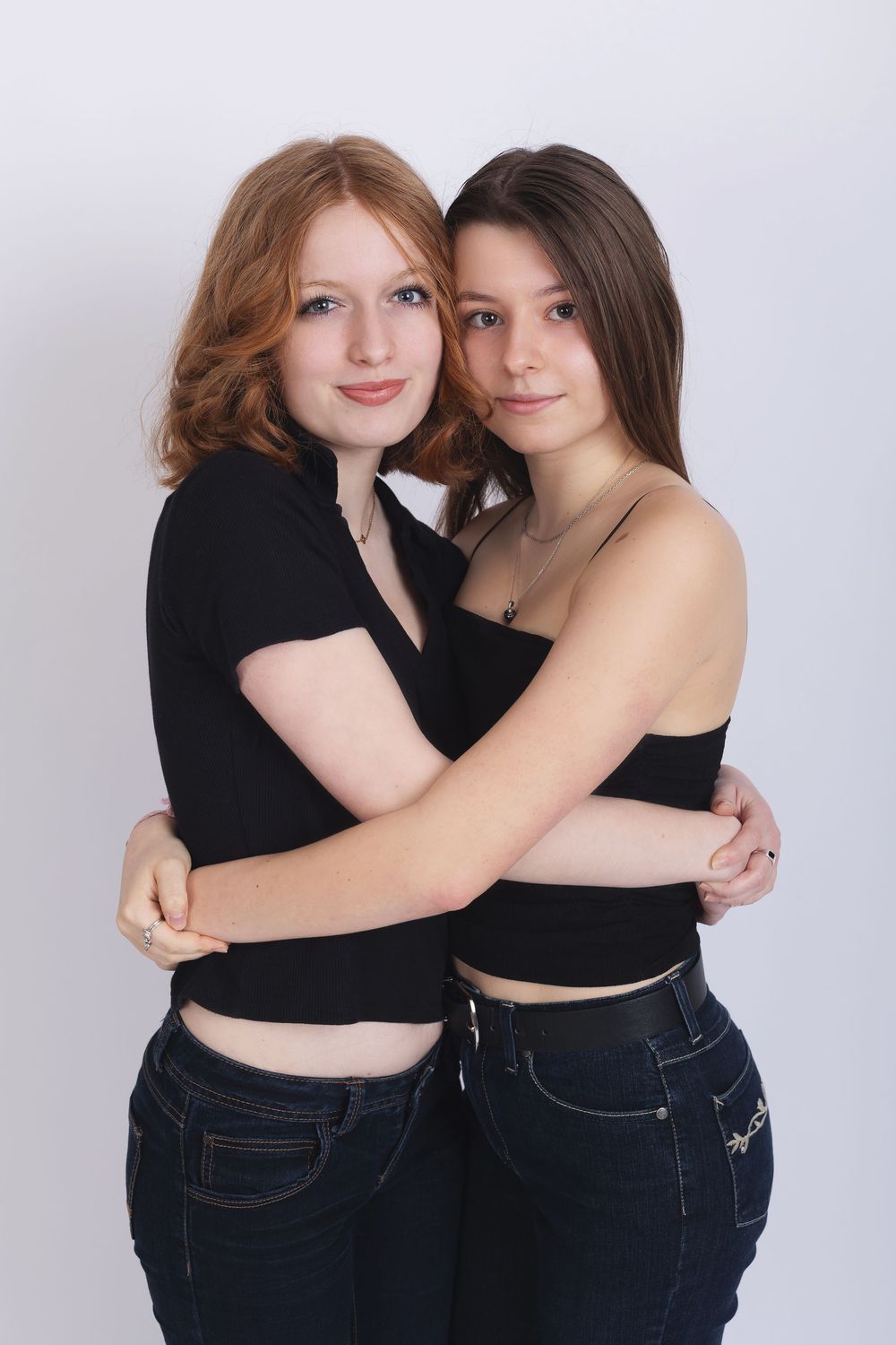 Two people in black tops and dark jeans embrace while posing together against a light background.