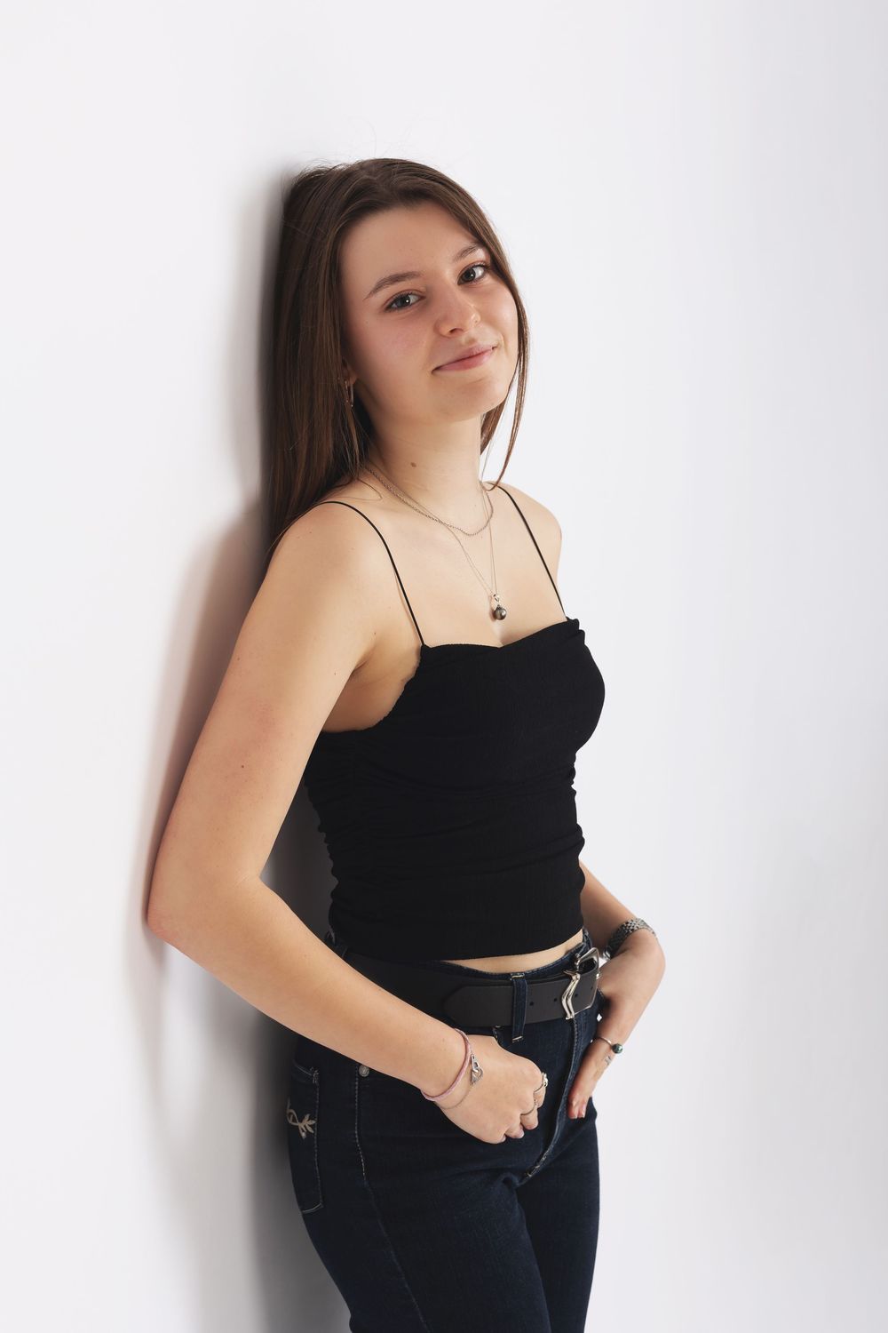 Portrait against white wall shows model in black crop top and dark jeans with delicate necklace and casual pose.