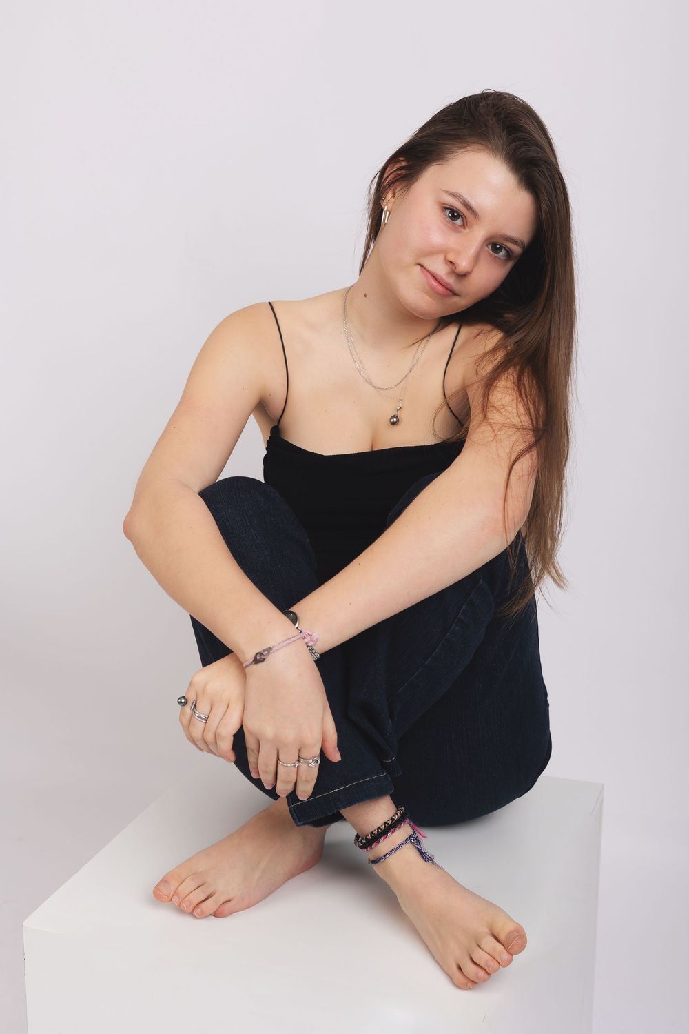 Portrait of a young woman in black clothing sitting casually on a white cube against a light background.