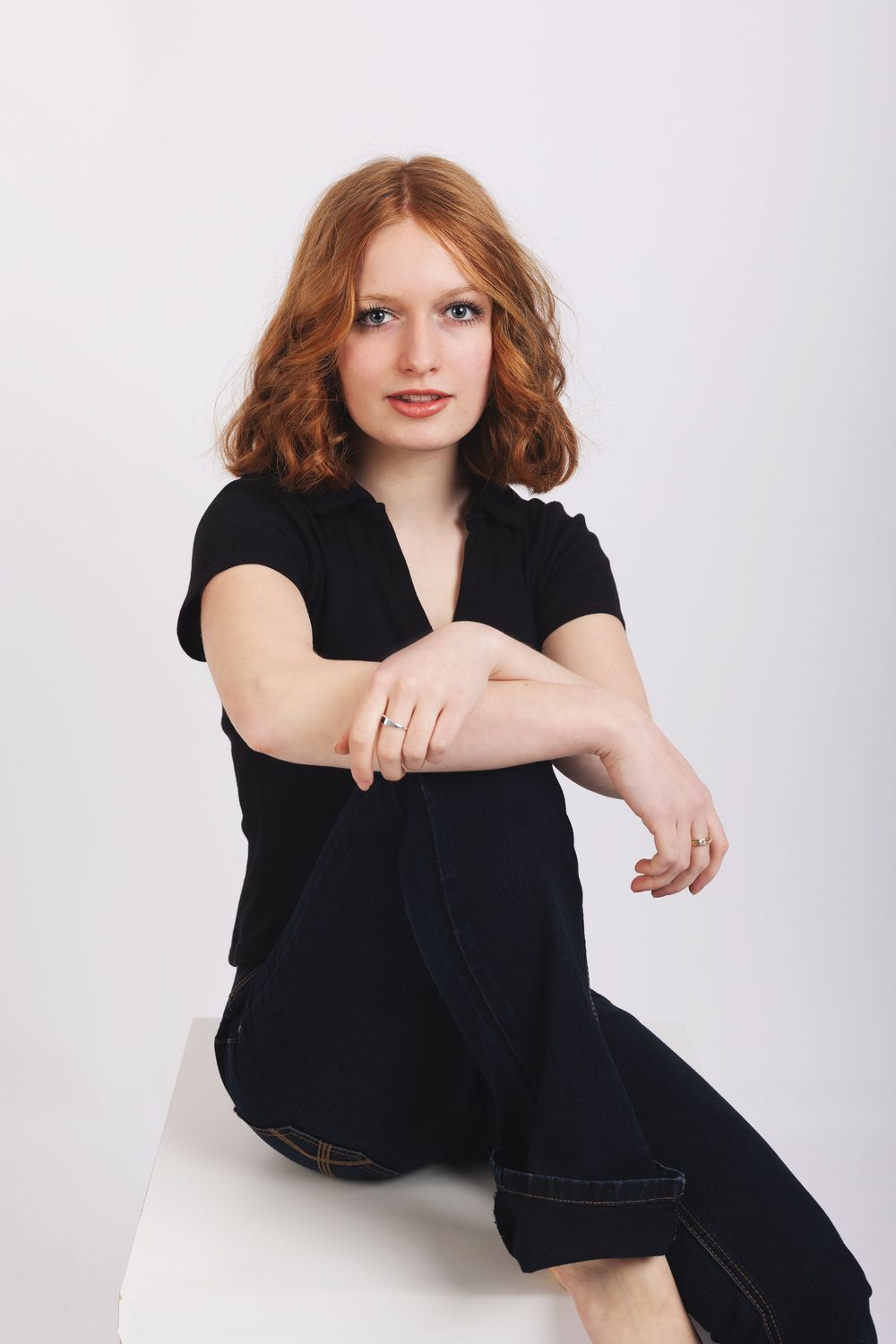 Portrait of someone with vibrant red wavy hair wearing a black top and jeans while sitting casually on a white surface.