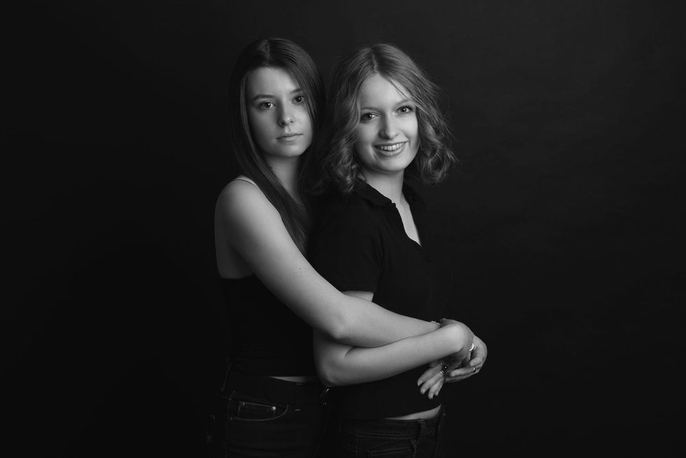 Striking black and white portrait of two people embracing while wearing dark clothing against dark background.