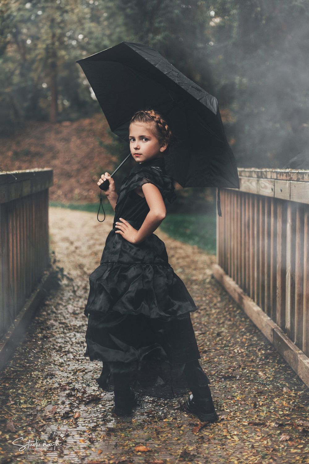 A small figure in a black dress holds a dark umbrella while standing on a misty wooden bridge covered in autumn leaves.