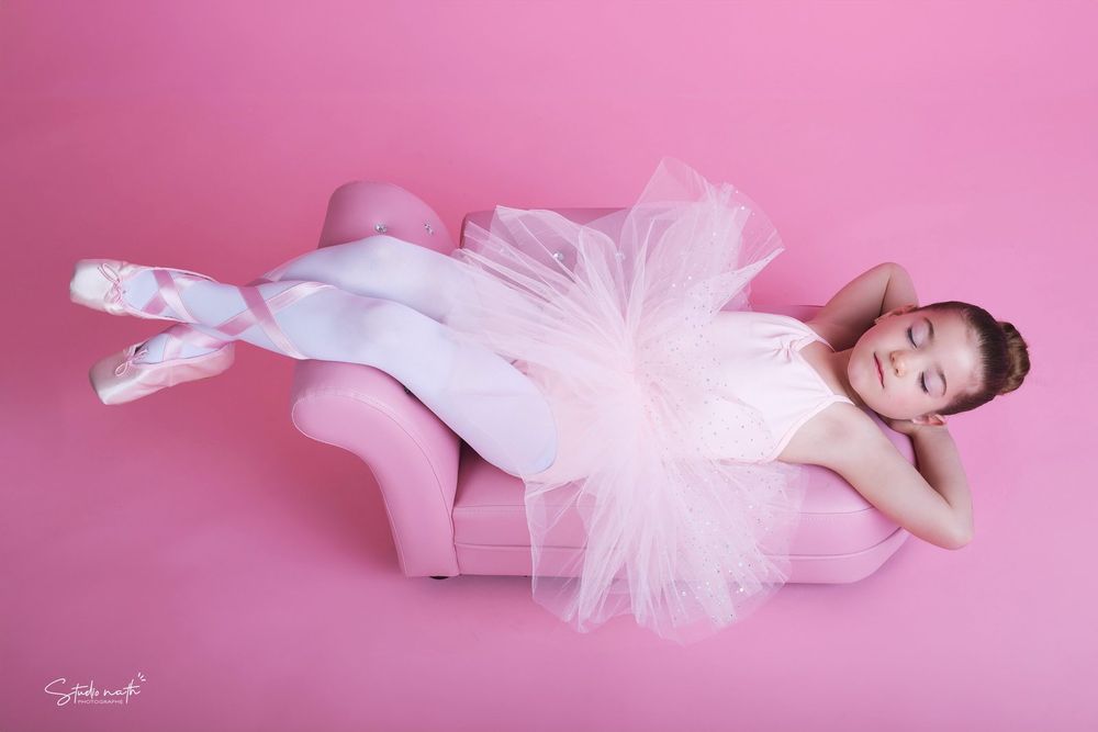 Dancer in white tulle dress posing gracefully on pink chaise lounge against rosy backdrop.