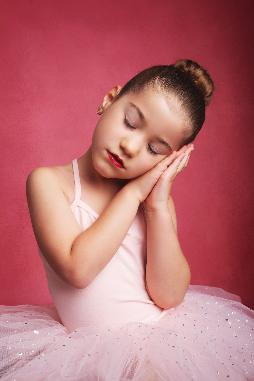 Young ballet dancer in pink tutu and leotard poses with eyes closed against a red background.