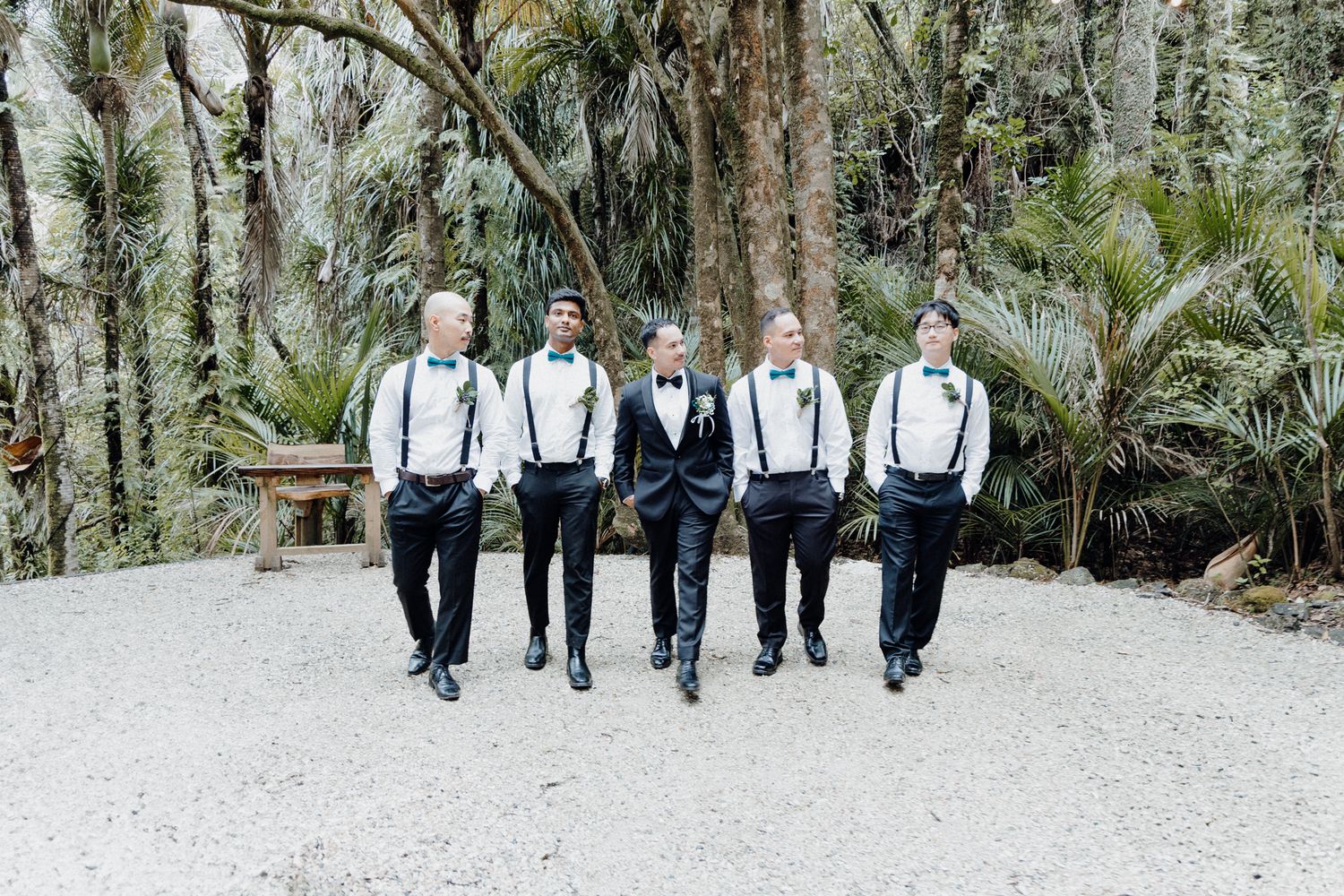 The groom's squad walk along a gravel path surrounded by tropical palm trees captured by Apertured Media Auckland Wedding Photographer.