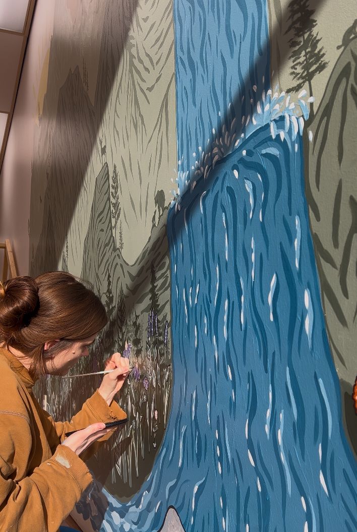 Shania Hirdler Mural Artist painting a large flower on a wall. Waterfall and mountains all around.