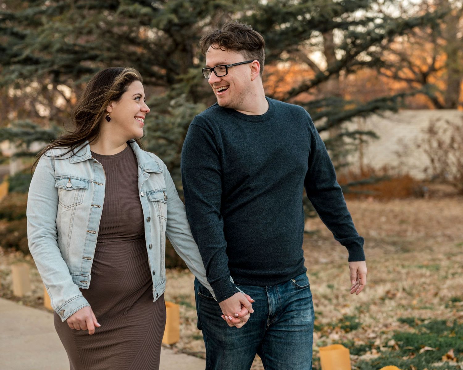 A couple walks hand in hand outdoors during golden hour, wearing casual fall attire against an autumn backdrop for engagement photo.