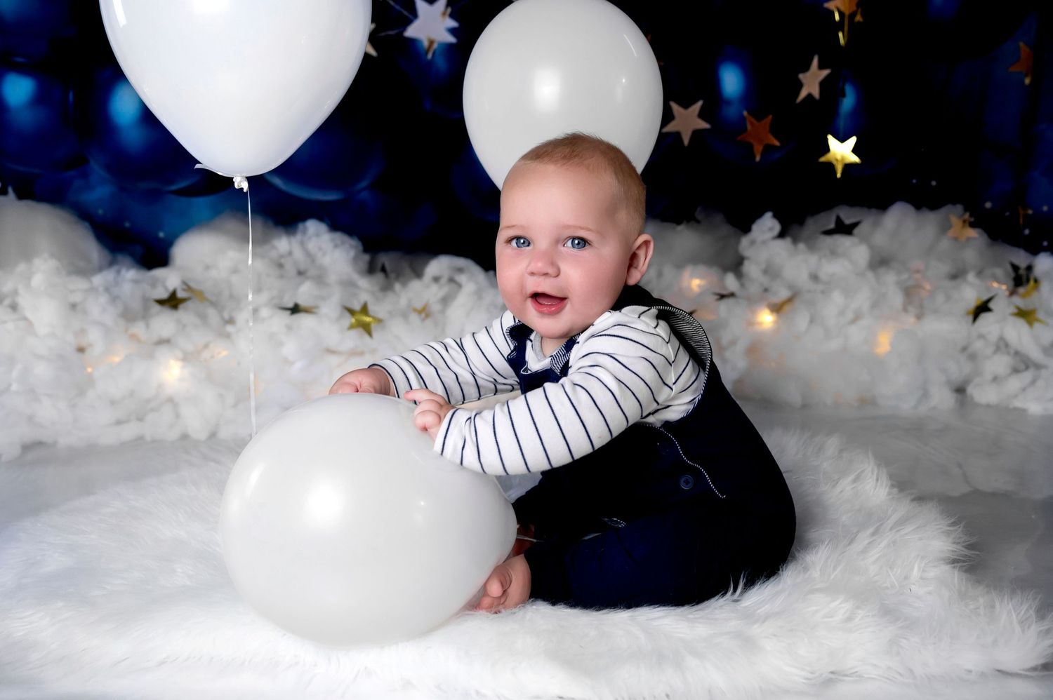 Baby in striped outfit playing with white balloons against blue starry backdrop.