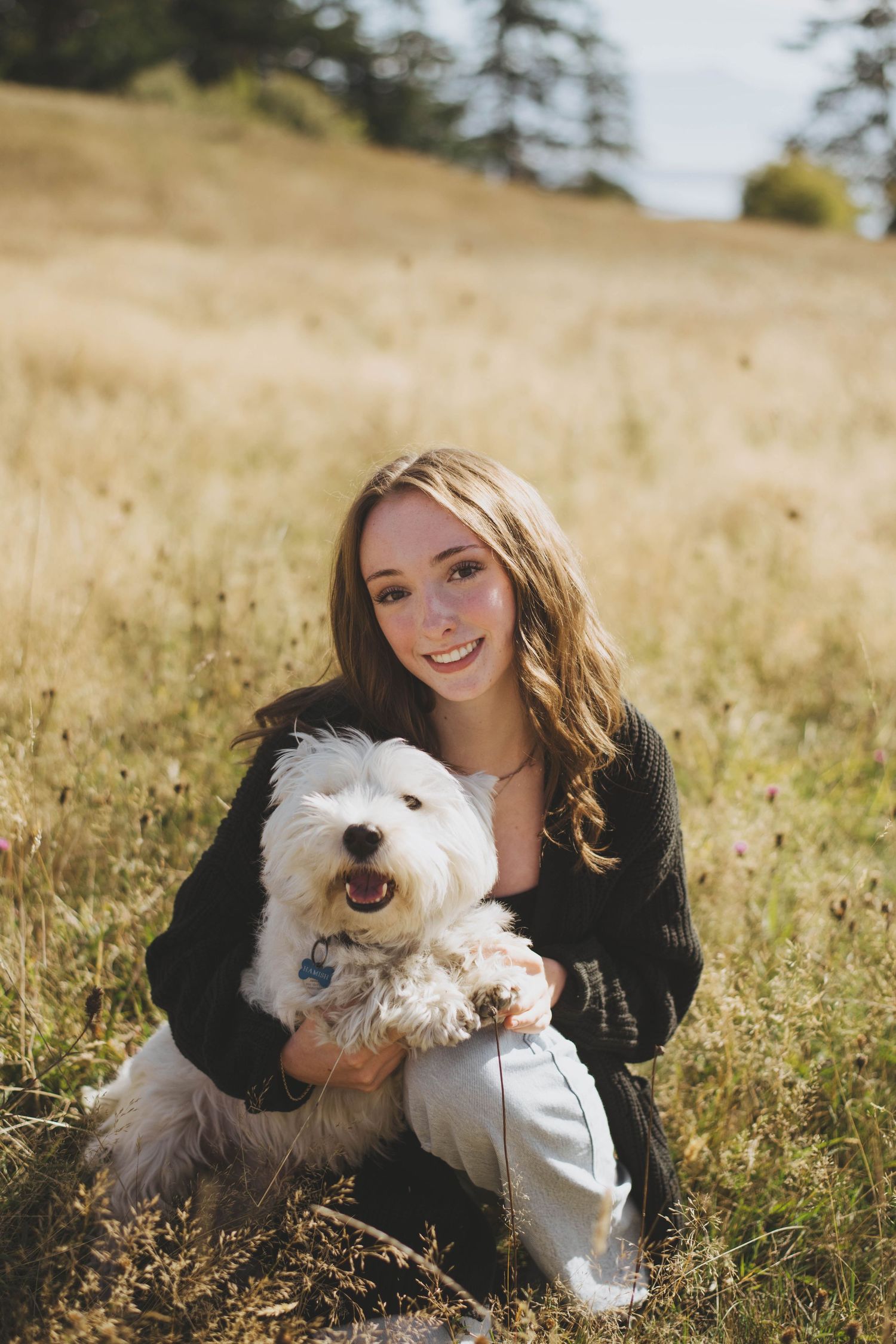 A young woman kneels in a grassy field, smiling warmly as she holds a happy, fluffy white dog in her arms.