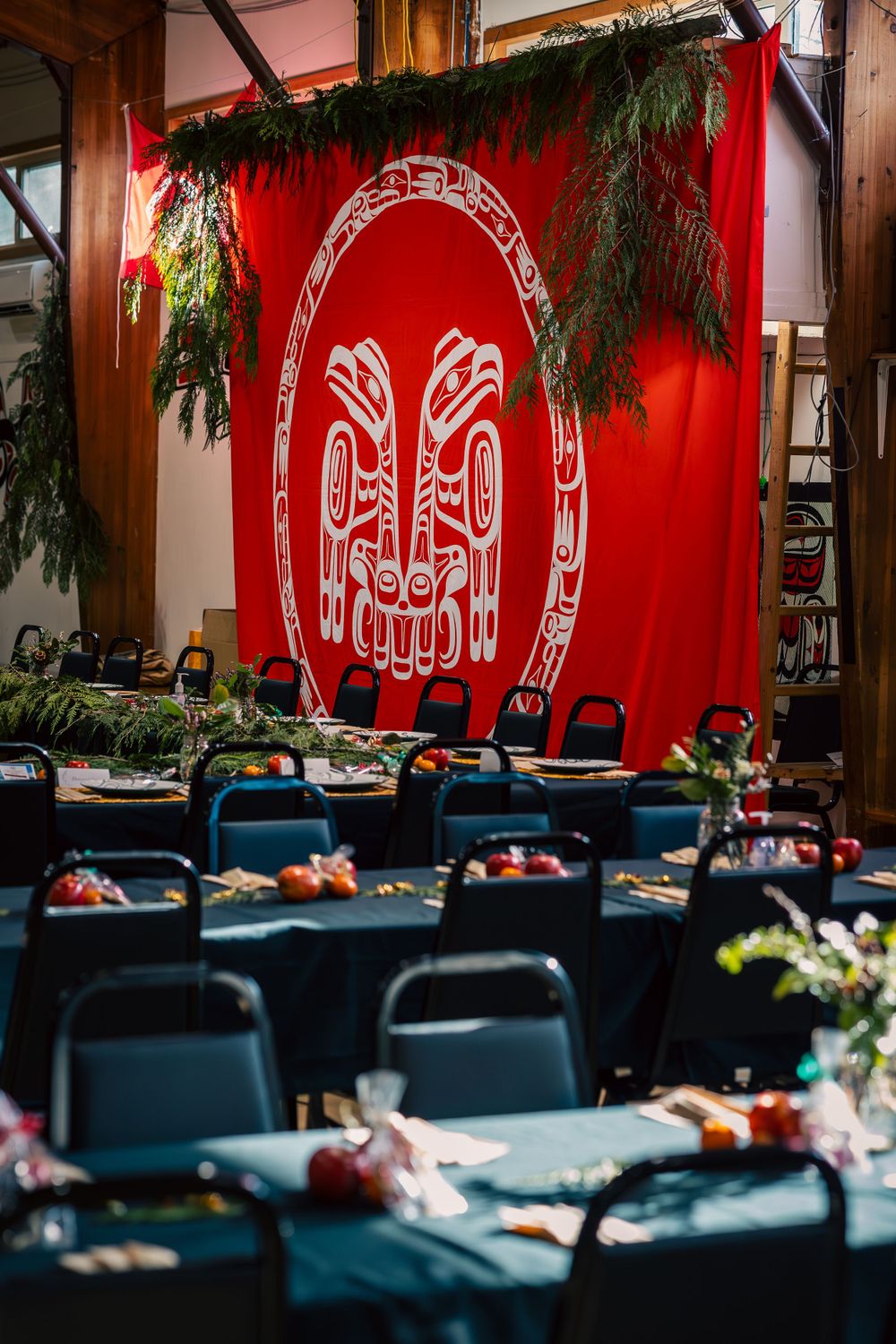 Event venue decorated with red banner featuring Haida art design and evergreen garlands over formal dining tables.