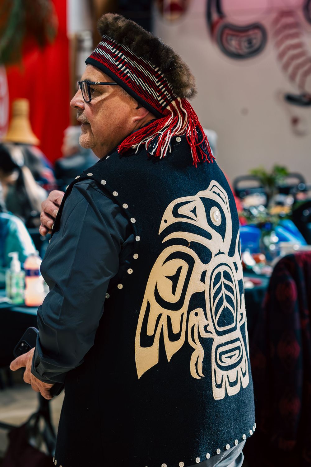 A person wearing a black jacket with Haida Indigenous art design and a winter hat at an indoor event.