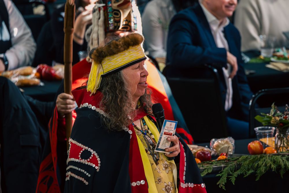 A person in traditional Haida regalia with a red blanket holds a ceremonial staff during a festive gathering.