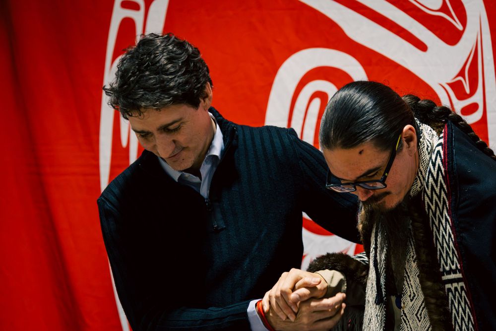 Two people look down at a mobile device in front of a red flag with indigenous artwork design in the background.