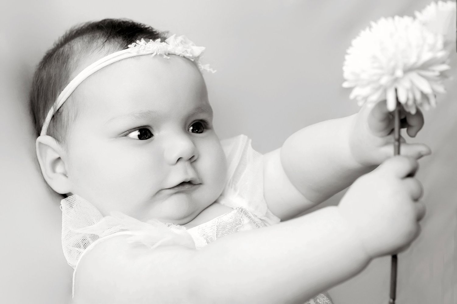 Black and white image of an infant reaching for a white flower while wearing a delicate headband.