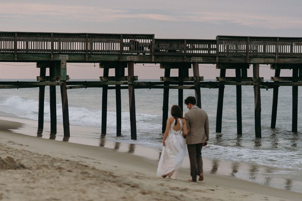 A couple walking along the beach beneath a wooden pier at sunset.