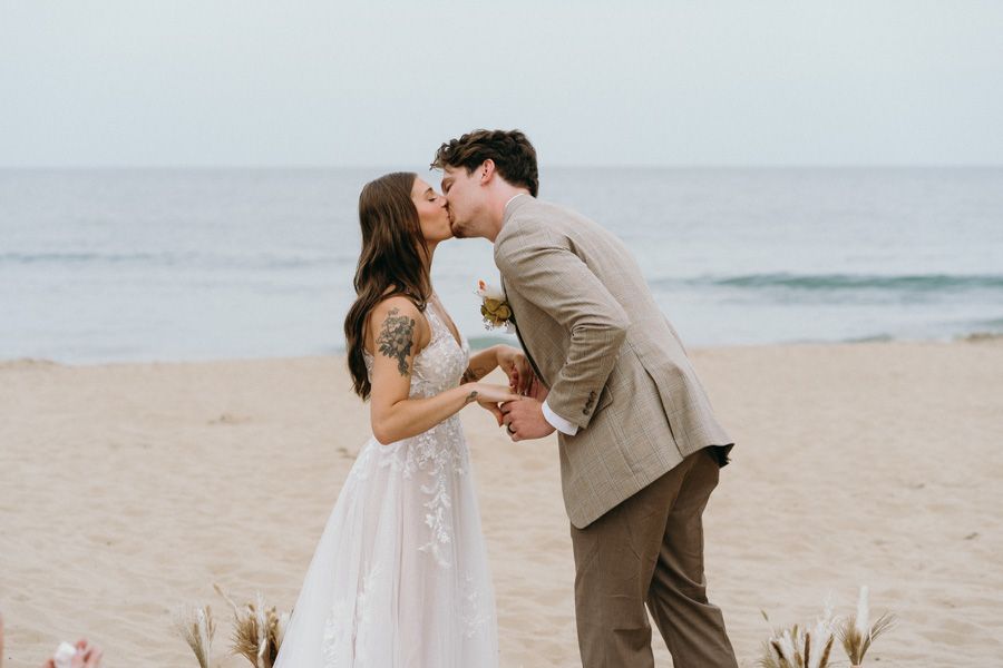 A newlywed couple sharing their first kiss on a beach with ocean waves in background.
