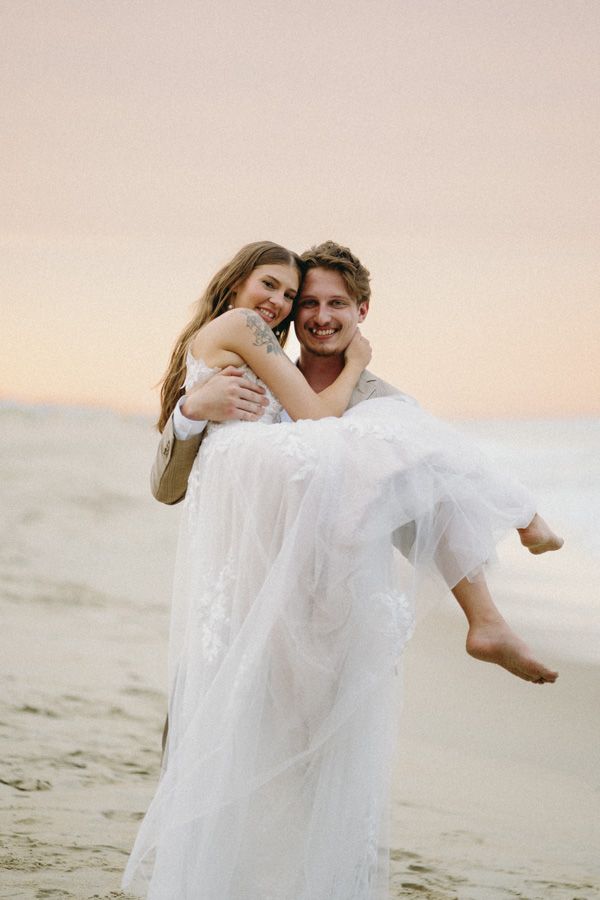 A groom carrying his bride on the beach at sunset with her dress flowing in the breeze.
