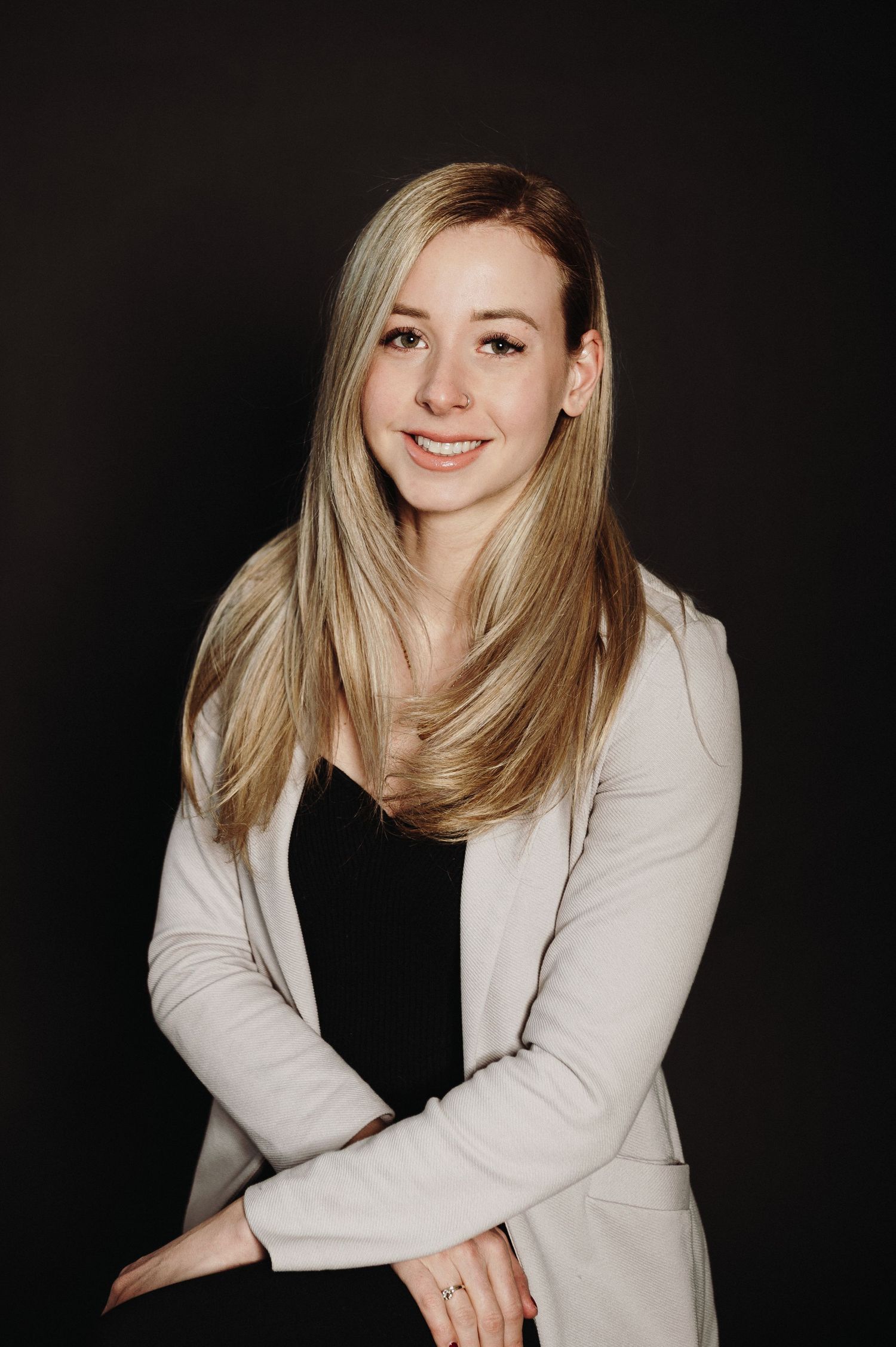 Professional portrait shot of someone posing in a white blazer and black dress against a dark background.