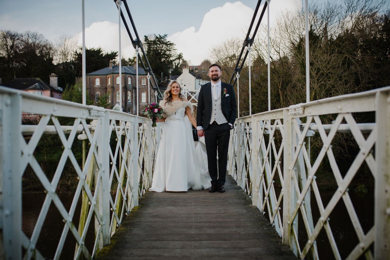 A newlywed couple walking together on a white latticed bridge during a scenic outdoor wedding ceremony.