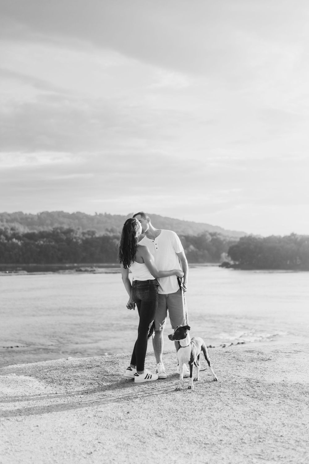 A couple crouches on a beach with their small dog while enjoying scenic waterfront views at sunset.