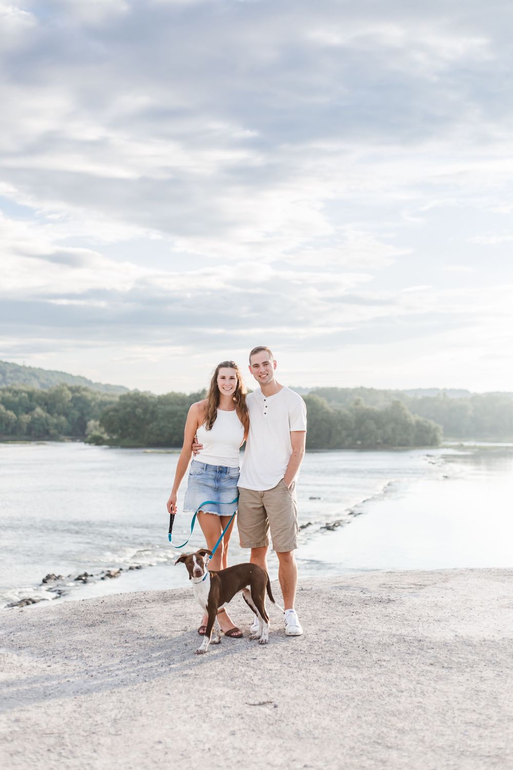 A couple and their brown dog enjoying a scenic walk along a waterfront path on a cloudy summer day.