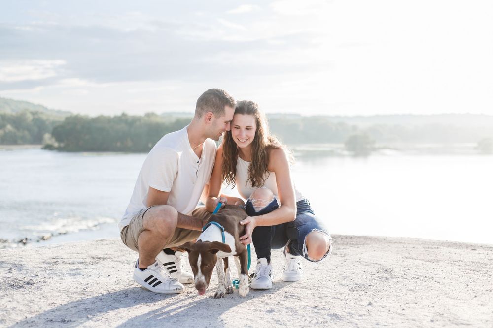 A couple crouches on a scenic beach overlook with their small brown and white dog during golden hour.