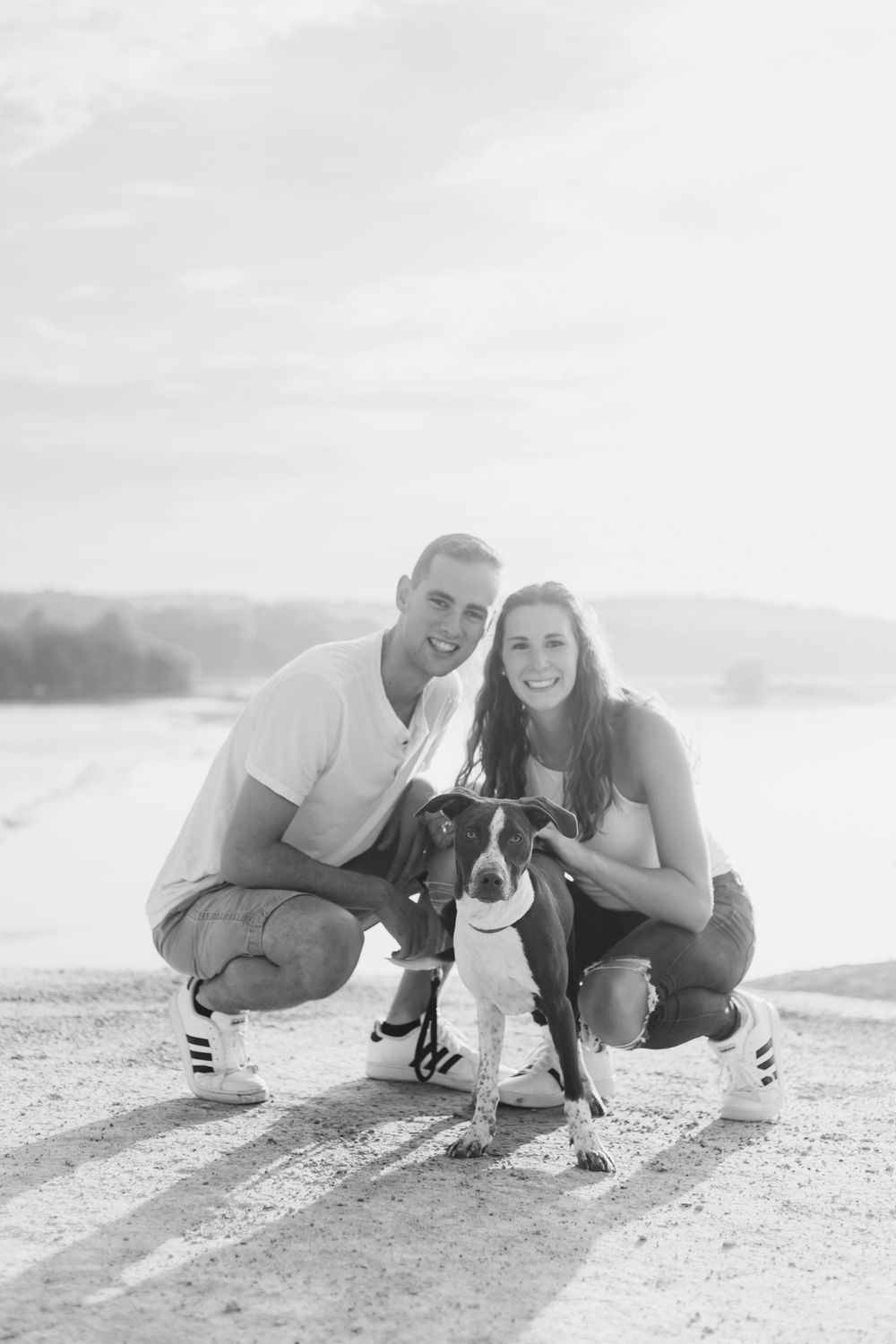 A couple and their small dog pose playfully together on a sunny beach overlook.
