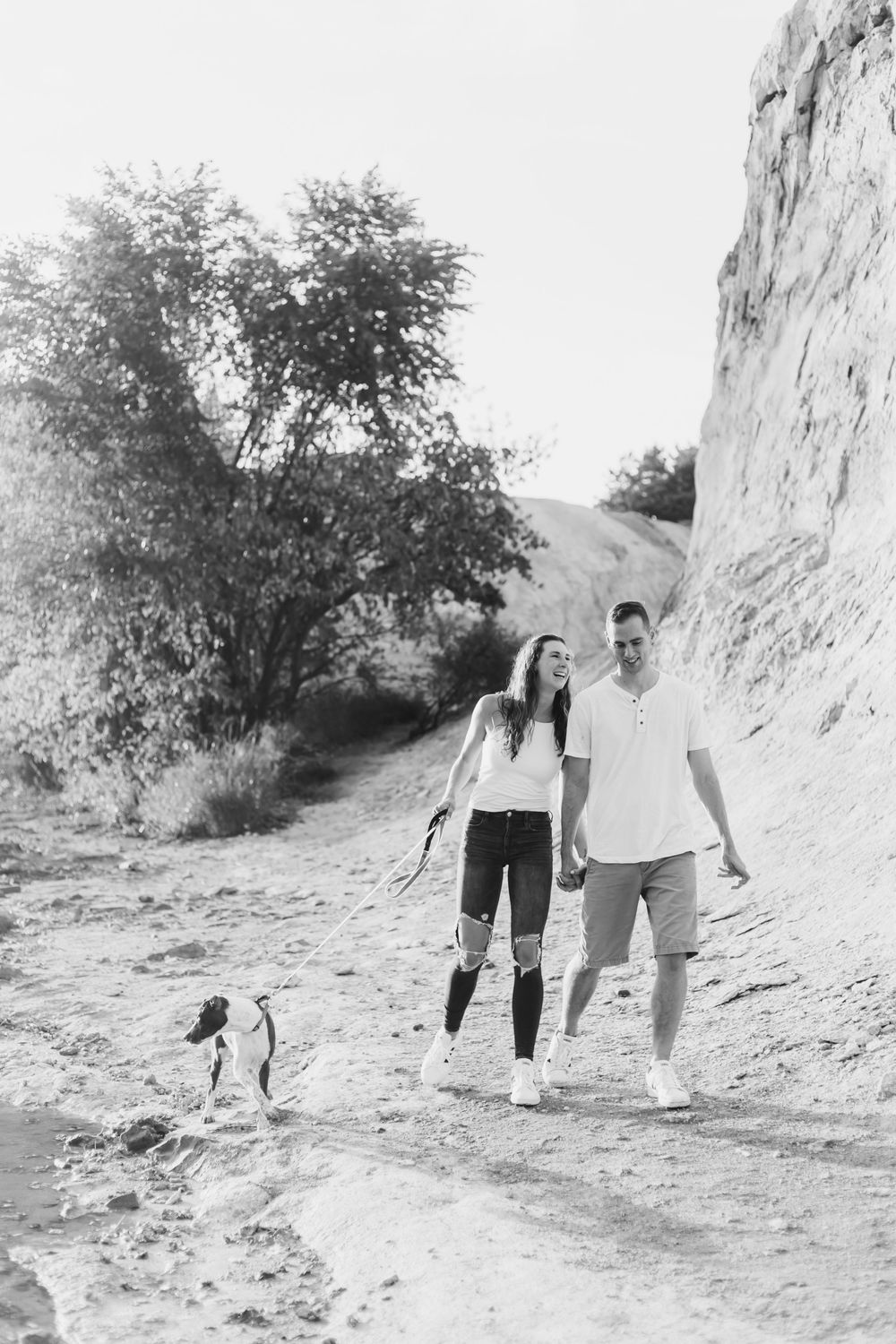 A couple walks hand in hand with their dog along a winding dirt path on a sunny afternoon.
