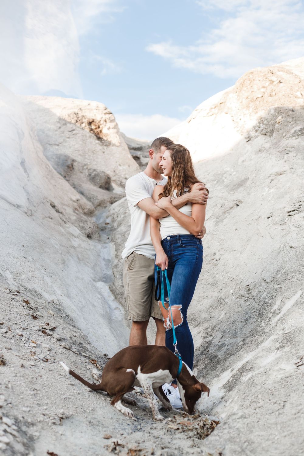 A couple embraces while standing in a white chalky canyon with their brown dog beside them on a sunny day.