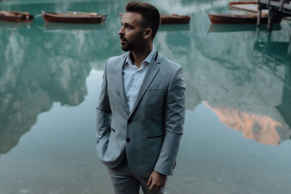 Professional wearing a gray suit stands by a serene waterfront setting with reflections in teal-tinted tones.
