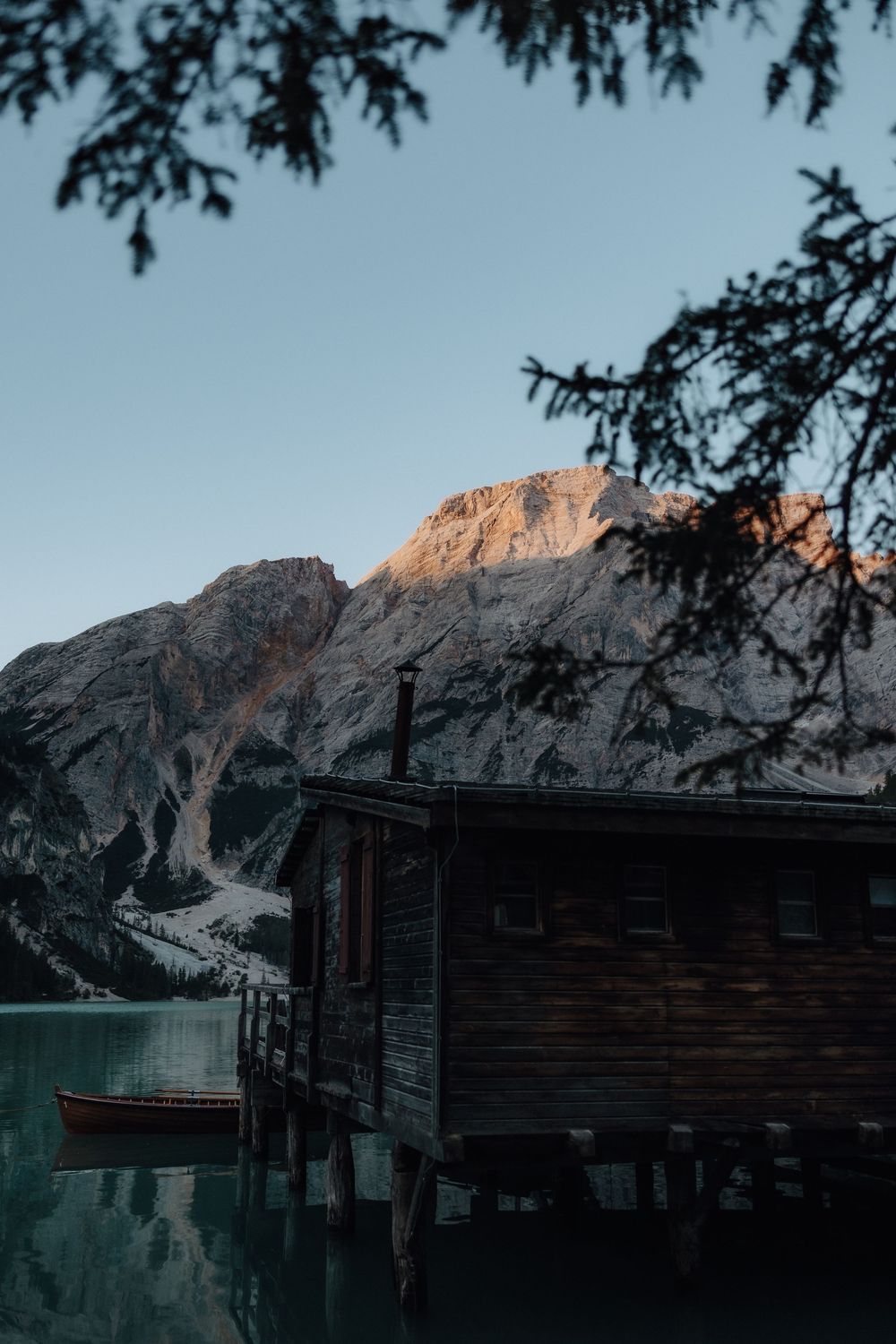 Lakeside mountain landscape with rustic wooden cabins silhouetted against snowy peaks at dusk.