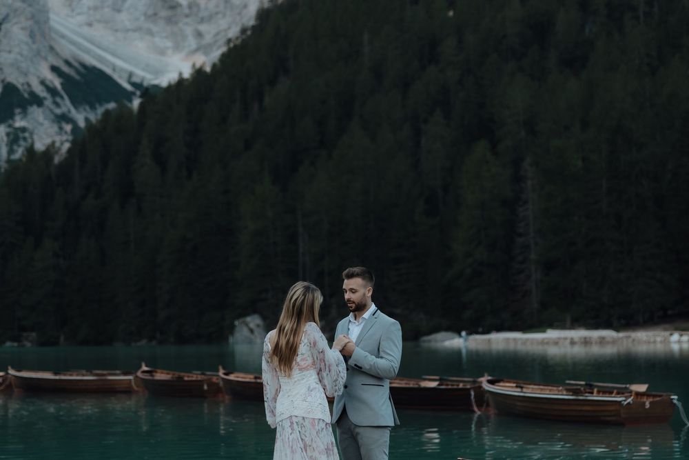 Outdoor mountain adventure photo session with couple hugging near lake surrounded by forest and snowy peaks.