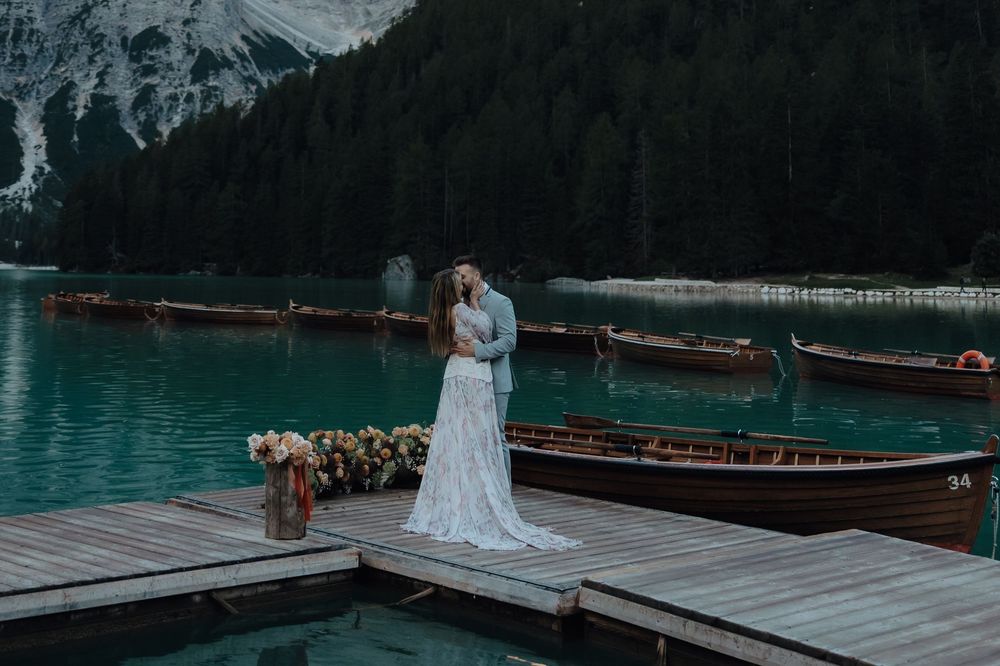 A couple embraces on a wooden dock overlooking turquoise alpine lake waters with snow-capped mountains in the background.
