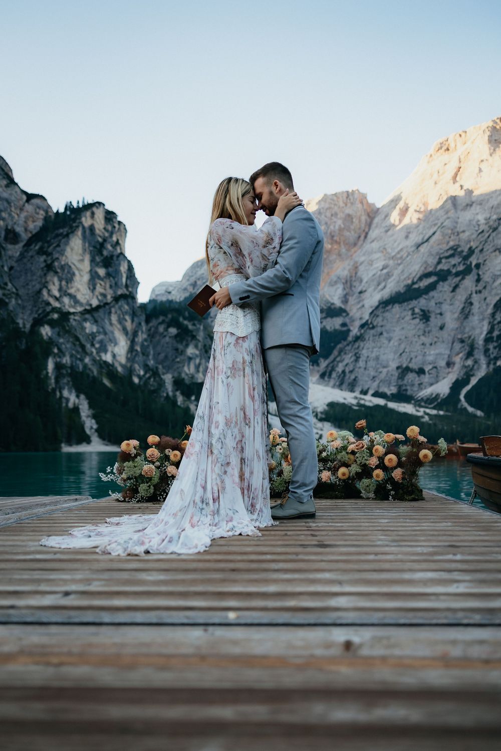 A romantic wedding couple embraces on a wooden pier with majestic mountains and crystal blue waters behind them.
