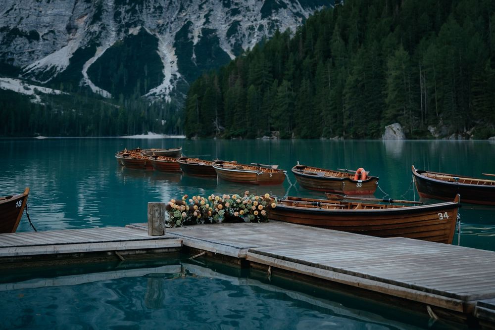 Wooden boats moored at a dock on a turquoise alpine lake surrounded by evergreen forests and mountain peaks.