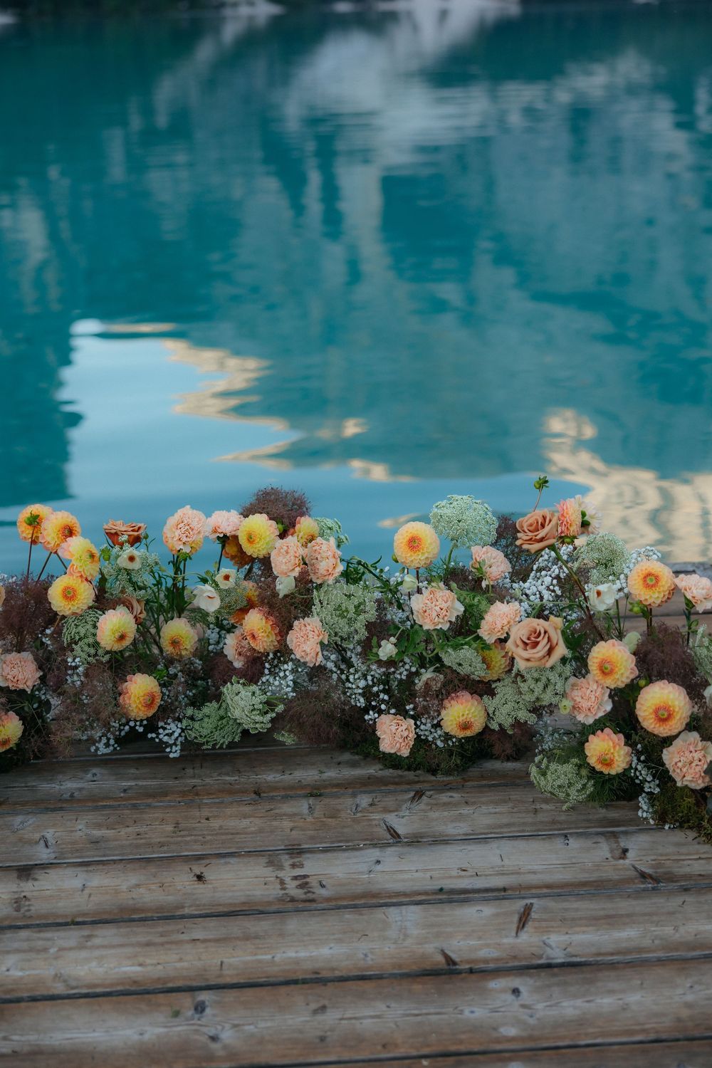 Delicate floral arrangement with peach and yellow blooms decorates a rustic wooden ledge against turquoise water reflections.
