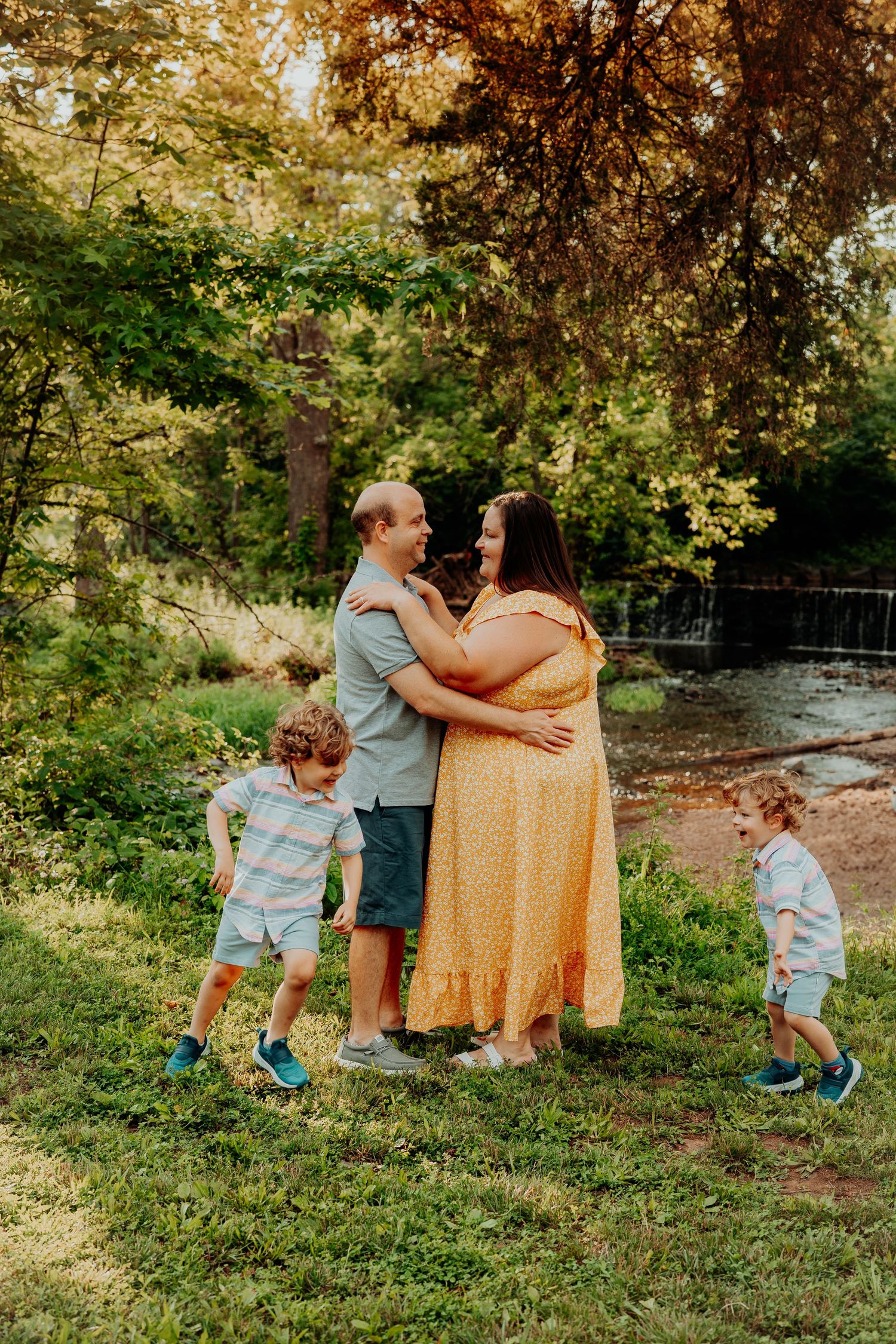 A family shares a tender moment together in a lush green park setting with a waterfall in the background on a sunny day.