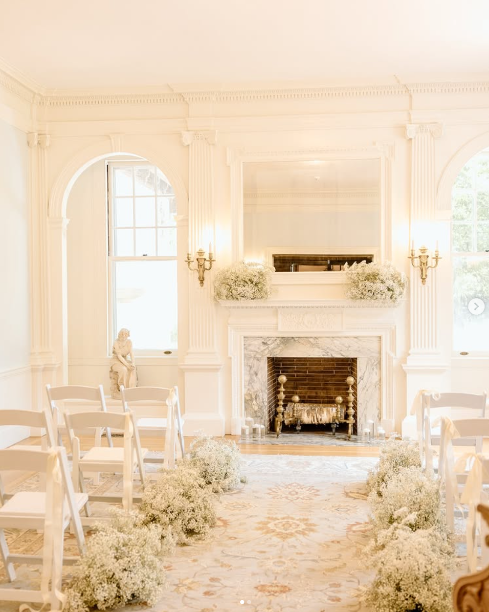 Elegant white wedding ceremony room with marble fireplace, arched window, and baby's breath floral arrangements along the aisle.