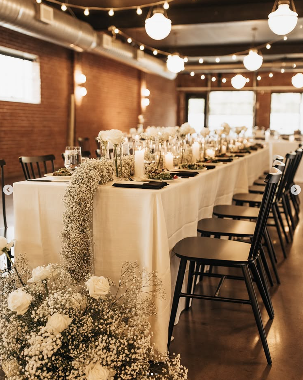Long banquet table with elegant white linens, delicate baby's breath floral arrangements, and black chairs in an industrial venue.