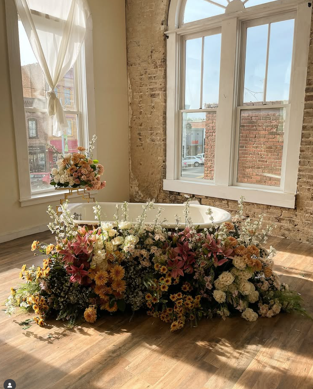 Abundant floral arrangement with wildflowers and roses sits in sunlit room with exposed brick walls and large windows.