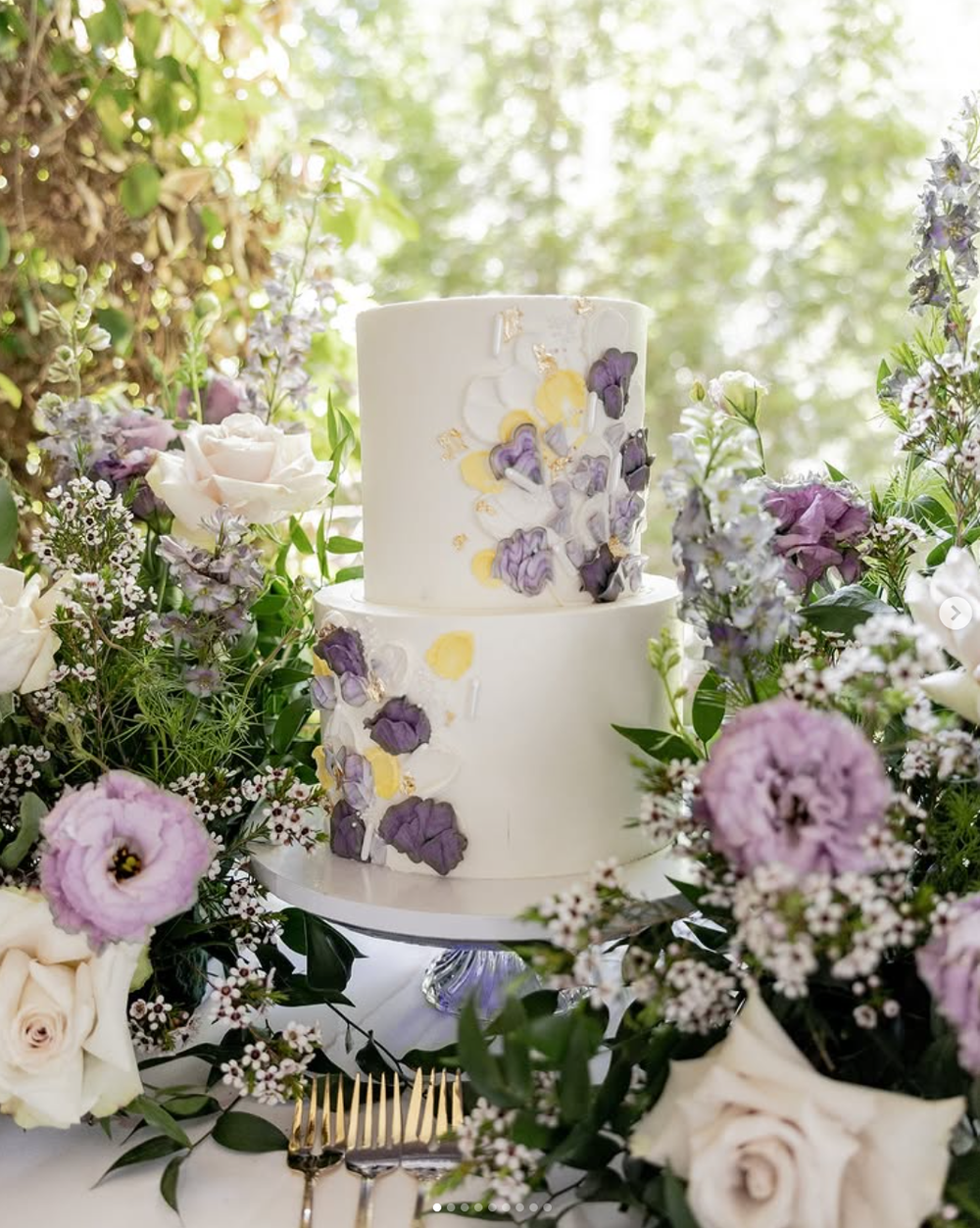 Elegant white wedding cake with purple and yellow floral decorations surrounded by fresh garden flowers and greenery.